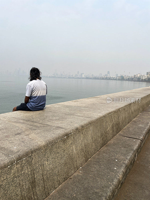 印度孟买，一名男子坐在海洋大道海堤上，混凝土防御，海岸管理，防止侵蚀，海水，c形长廊，装饰艺术建筑和摩天大楼排列在Girgaon Chowpatty公共海滩的后视图
